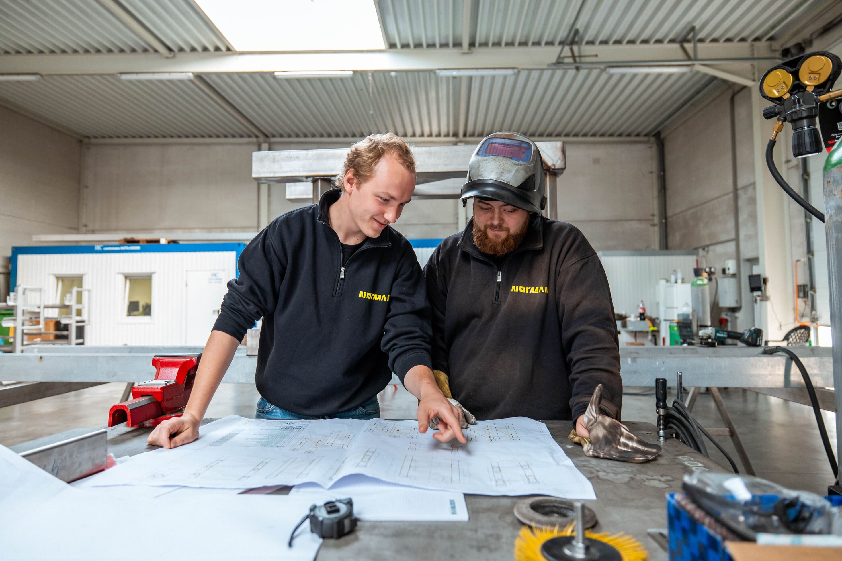 Twee N&oslash;rmannen bekijken samen een technische tekening in het atelier.